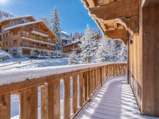 Experience the charm of a winter wonderland from this stunning wooden balcony, offering breathtaking views of snow-covered chalets and majestic pine trees.