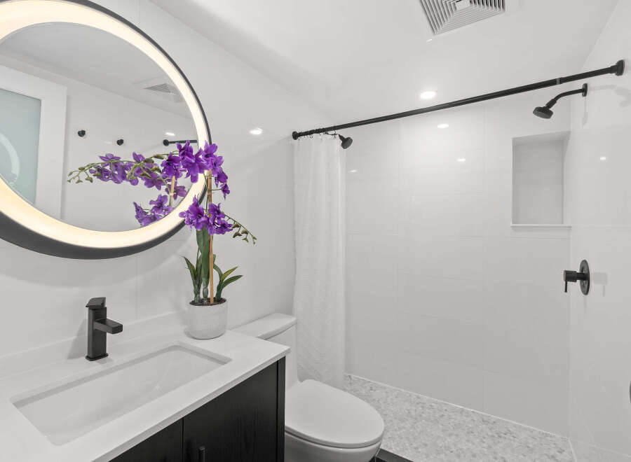Step into a modern bathroom oasis! This stunning space features sleek white tiles, a stylish black vanity, and a beautiful round mirror with integrated lighting.