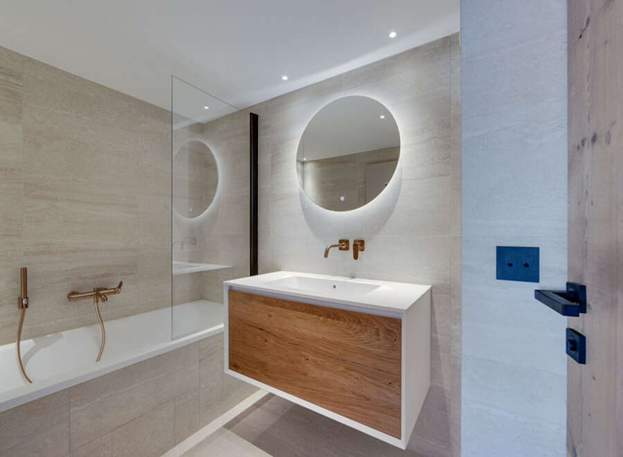 This modern bathroom boasts a sleek design with a wooden vanity and a round mirror with integrated lighting.