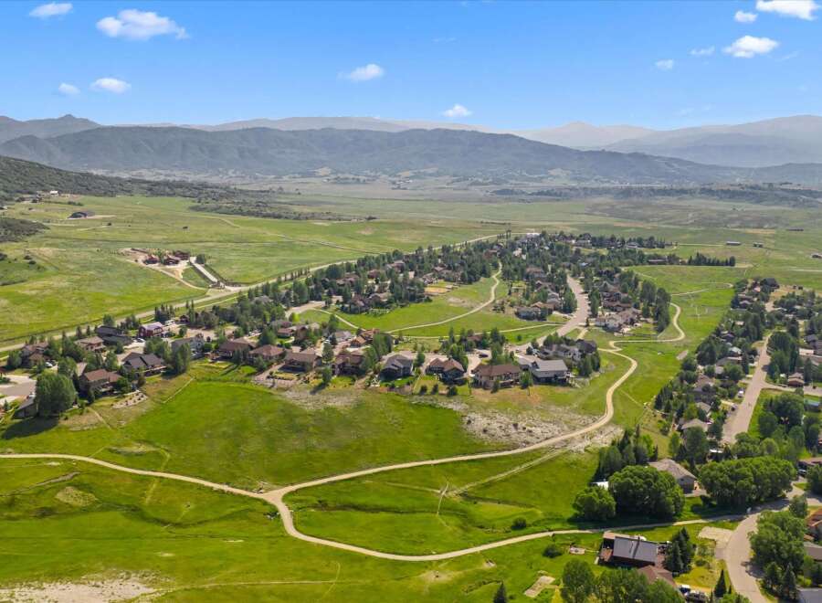Discover this stunning aerial view of a picturesque suburban neighborhood nestled amidst rolling green hills and majestic mountains.