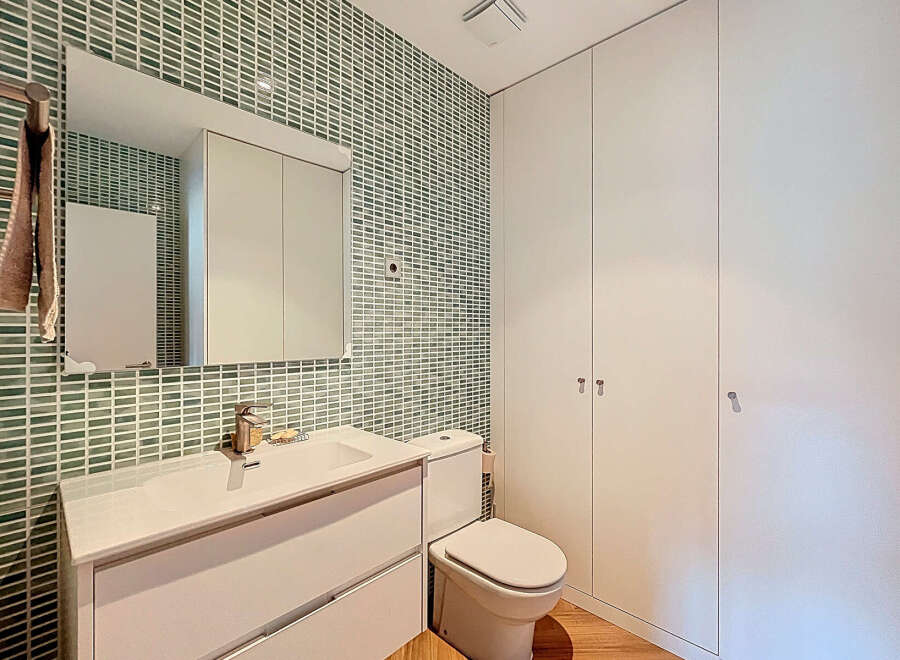 This modern bathroom features a sleek white vanity and a matching toilet, complemented by a stylish green tile backsplash.