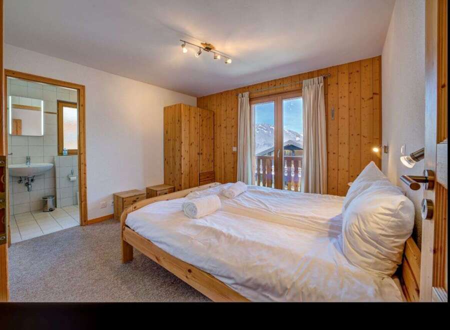 Experience cozy mountain living in this charming bedroom featuring a comfortable bed, ample wooden wardrobe, and a private balcony with stunning alpine views.