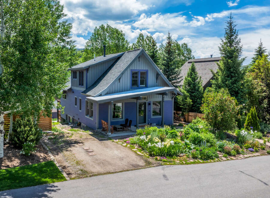 Charming purple home nestled among mature trees, featuring a welcoming front porch with seating and a vibrant garden bursting with colorful flowers.