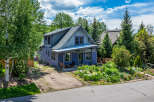 Charming purple home nestled among mature trees, featuring a welcoming front porch with seating and a vibrant garden bursting with colorful flowers.