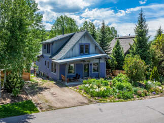 Charming purple home nestled among mature trees, featuring a welcoming front porch with seating and a vibrant garden bursting with colorful flowers.