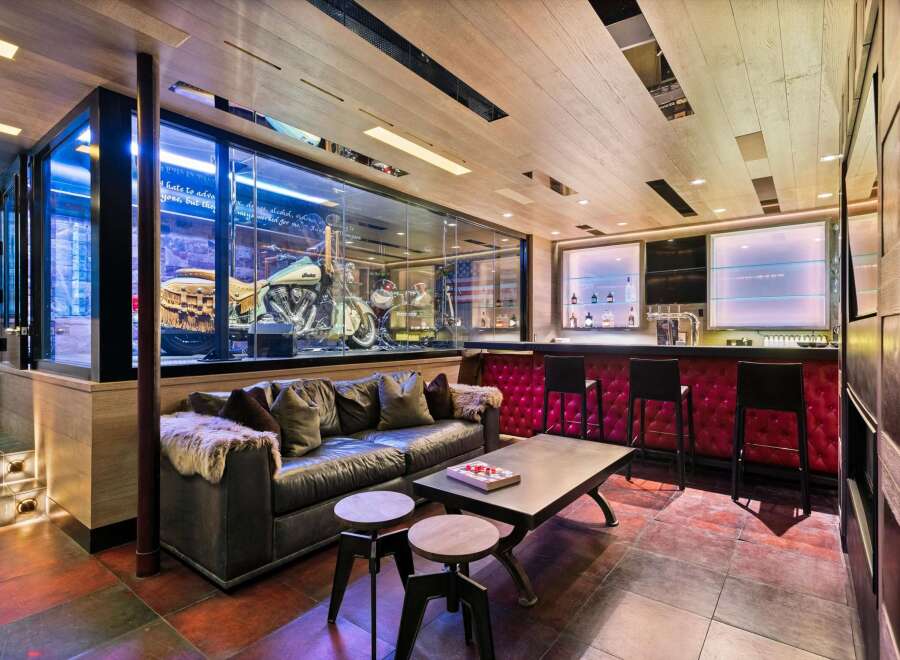 Experience ultimate luxury in this custom-designed entertainment space, featuring a stunning motorcycle display encased in glass, a plush leather sofa, and a fully equipped bar with plush red seating.
