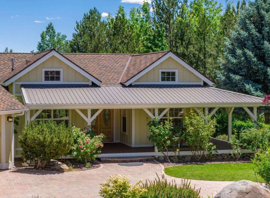 Charming single-family home featuring a welcoming covered porch with a metal roof and decorative white beams, perfect for enjoying the surrounding mature trees and lush landscaping.