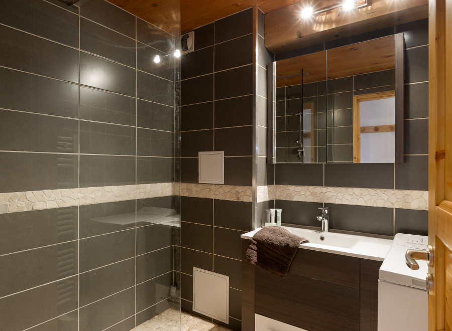 This modern bathroom boasts dark gray tiled walls, a glass shower enclosure, and a stylish dark wood vanity with a white sink.