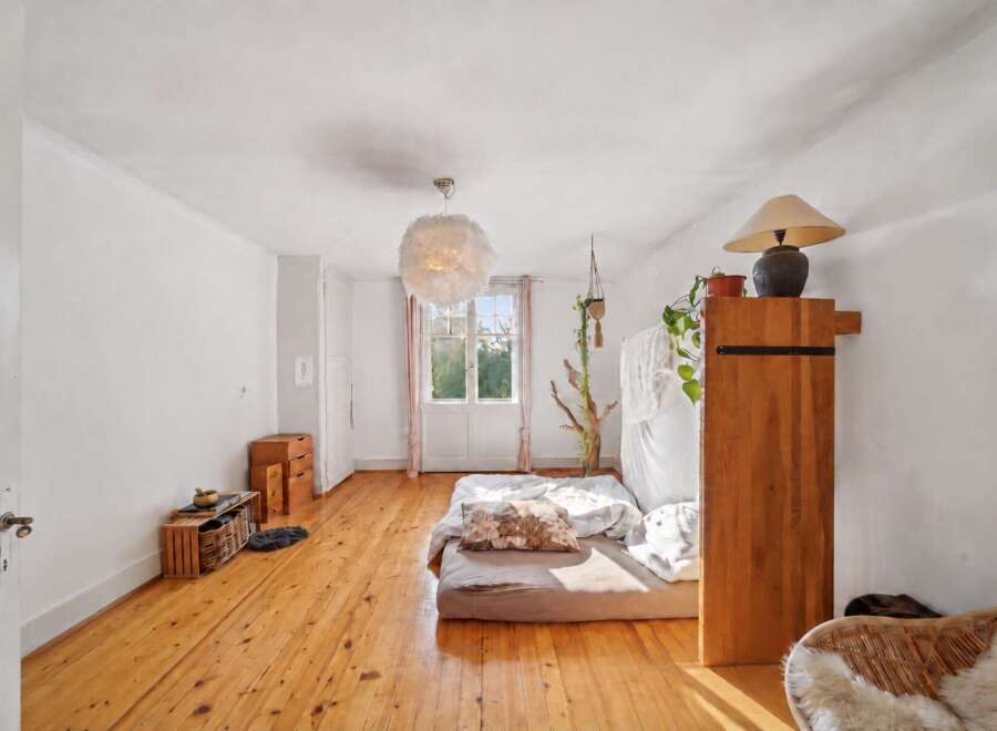 This bright and airy bedroom features a low-profile bed with a decorative pillow, set against a backdrop of warm, natural wood floors and white walls.