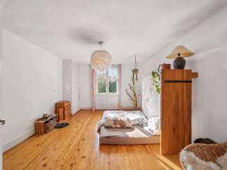 This bright and airy bedroom features a low-profile bed with a decorative pillow, set against a backdrop of warm, natural wood floors and white walls.