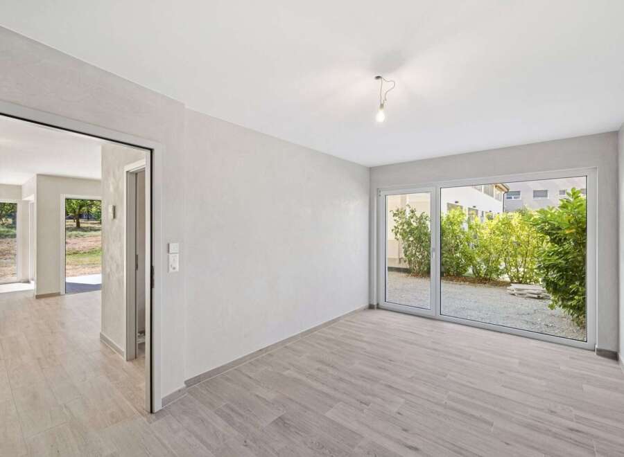 Discover modern living in this bright and airy space, featuring sleek light wood-look flooring and expansive glass doors that open to a lush green courtyard.