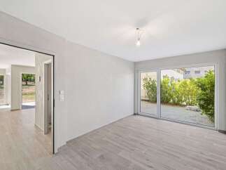 Discover modern living in this bright and airy space, featuring sleek light wood-look flooring and expansive glass doors that open to a lush green courtyard.