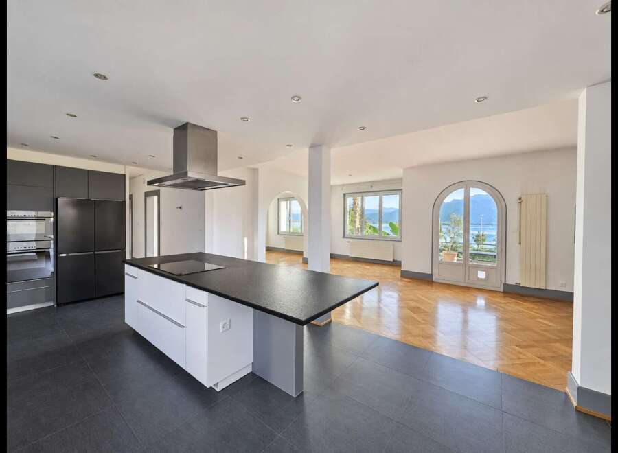 Experience modern living in this spacious open-plan kitchen and living area, featuring sleek dark cabinetry, a central island with a cooktop, and a stunning view of the lake and mountains through arched doorways.