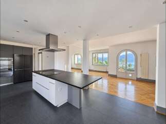 Experience modern living in this spacious open-plan kitchen and living area, featuring sleek dark cabinetry, a central island with a cooktop, and a stunning view of the lake and mountains through arched doorways.