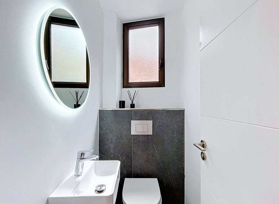 This modern bathroom features a sleek oval mirror with integrated LED lighting, a minimalist white vessel sink with a chrome faucet, and a wall-mounted toilet with a contemporary flush plate.
