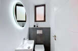 This modern bathroom features a sleek oval mirror with integrated LED lighting, a minimalist white vessel sink with a chrome faucet, and a wall-mounted toilet with a contemporary flush plate.