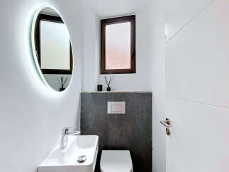 This modern bathroom features a sleek oval mirror with integrated LED lighting, a minimalist white vessel sink with a chrome faucet, and a wall-mounted toilet with a contemporary flush plate.