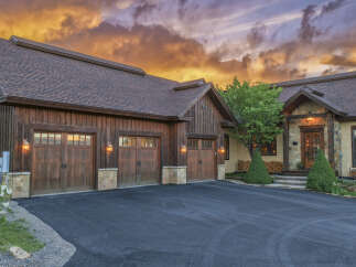 Experience luxury mountain living in this stunning timber-frame home, featuring a spacious four-car garage and a dramatic sunset sky.