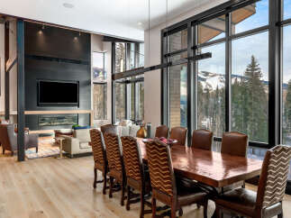 Experience luxury mountain living in this stunning dining and living area, featuring a modern fireplace and expansive windows with breathtaking views of snow-capped peaks and evergreen forests.