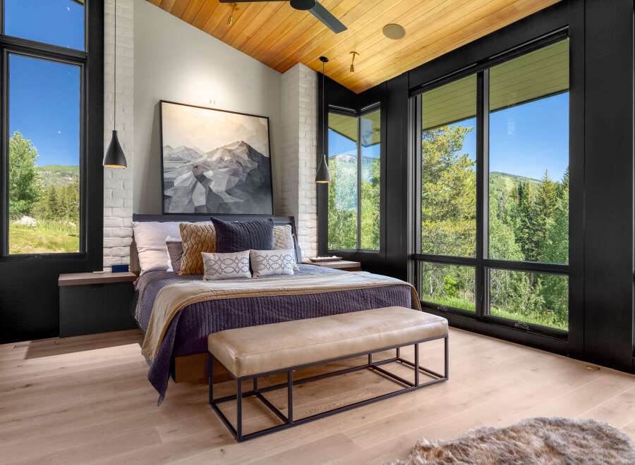 Experience luxury mountain living in this stunning bedroom featuring a vaulted wood-paneled ceiling and expansive windows that frame breathtaking forest and mountain views.