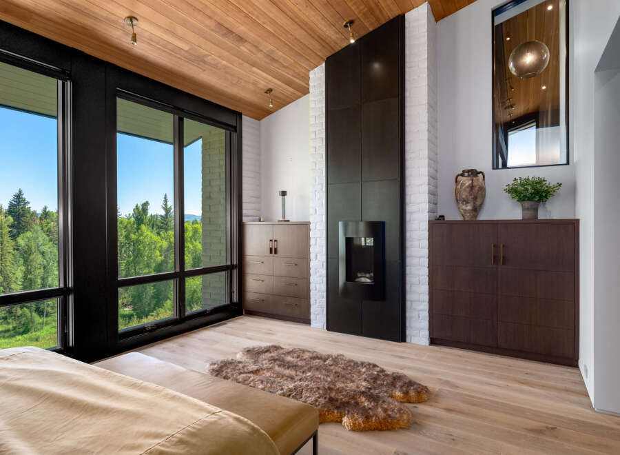 Experience luxury living with this stunning bedroom featuring a modern fireplace and expansive windows offering breathtaking forest views.
