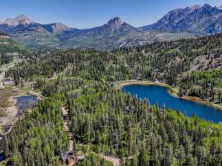 Discover your dream mountain retreat with this stunning aerial view showcasing a serene blue lake nestled amidst lush green forests and majestic peaks.