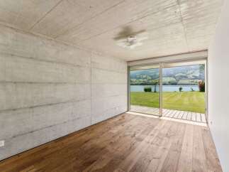 Experience modern living with this stunning concrete and wood interior, featuring a breathtaking view of a serene lake and rolling hills through expansive glass doors.
