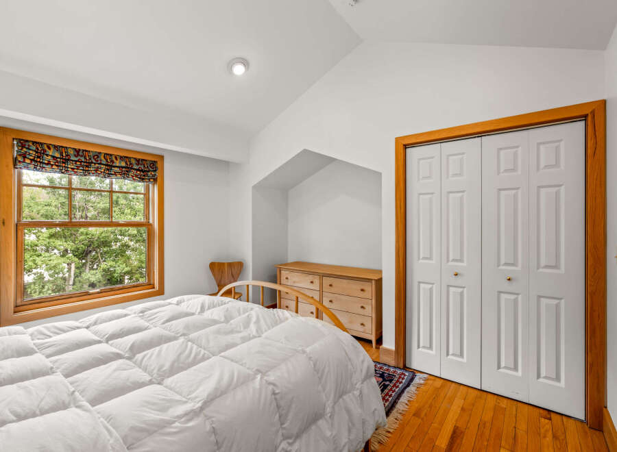 This charming bedroom features a large window with a colorful roman shade, offering a view of lush greenery.