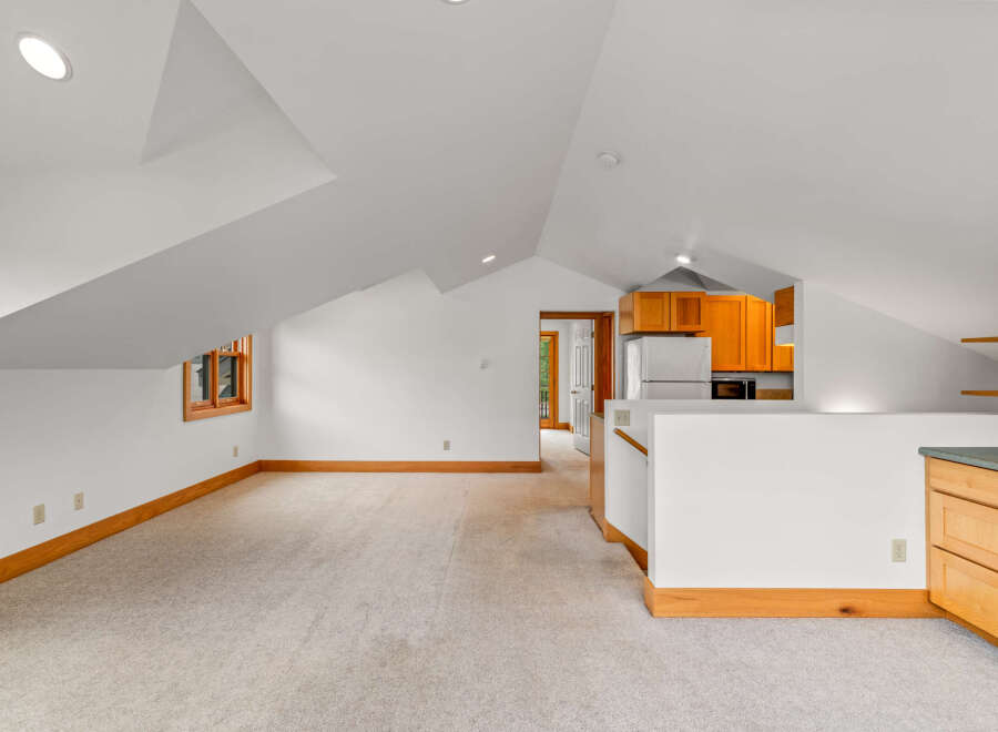 Discover this charming attic space featuring a cozy living area with plush carpeting and a convenient kitchenette equipped with wooden cabinetry and a refrigerator.