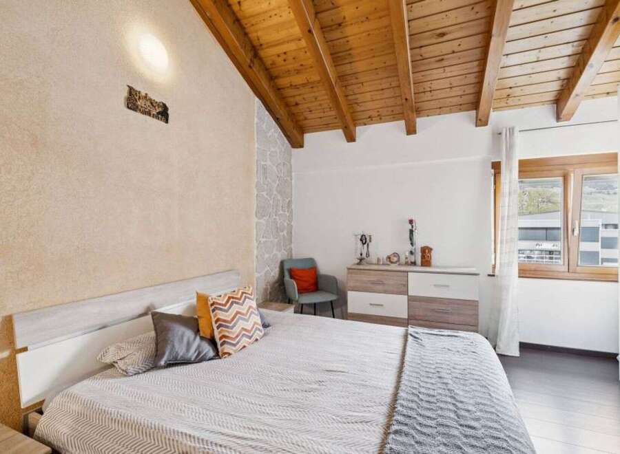 This charming bedroom features a cozy bed with patterned pillows and a soft throw, complemented by a rustic stone accent wall and a warm wooden ceiling.