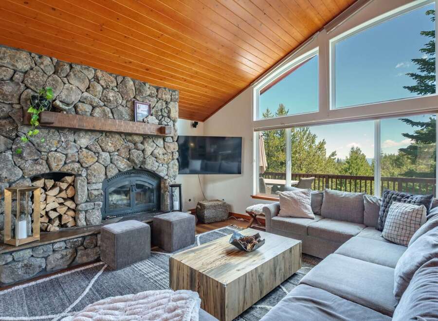 Experience breathtaking mountain views from this stunning living room, featuring a grand stone fireplace and a vaulted wood-paneled ceiling.