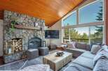 Experience breathtaking mountain views from this stunning living room, featuring a grand stone fireplace and a vaulted wood-paneled ceiling.