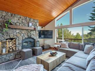 Experience breathtaking mountain views from this stunning living room, featuring a grand stone fireplace and a vaulted wood-paneled ceiling.