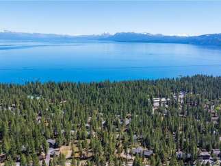 Experience the ultimate lakeside living with this stunning aerial view of homes nestled among tall pine trees, offering breathtaking vistas of a vast, blue lake and distant snow-capped mountains.
