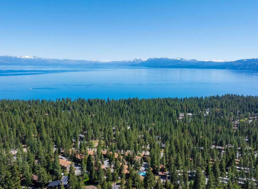Experience the ultimate lakeside living with this stunning aerial view of homes nestled amidst a dense pine forest, offering breathtaking vistas of a vast, shimmering lake and distant snow-capped mountains.