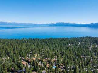 Experience the ultimate lakeside living with this stunning aerial view of homes nestled amidst a dense pine forest, offering breathtaking vistas of a vast, shimmering lake and distant snow-capped mountains.