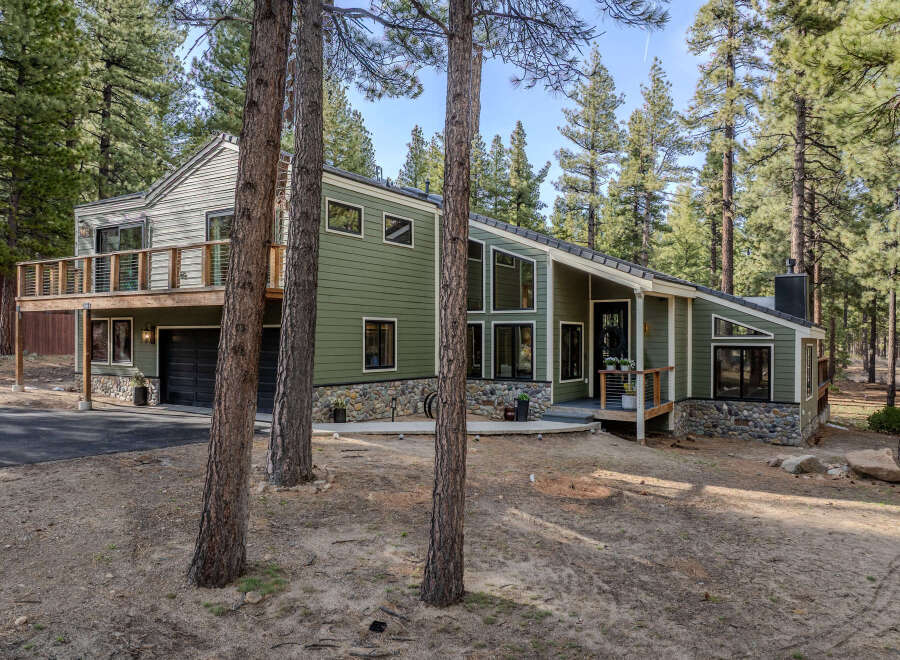 Nestled among tall pines, this modern mountain home boasts a striking green exterior with stone accents and a multi-level deck perfect for enjoying the serene surroundings.