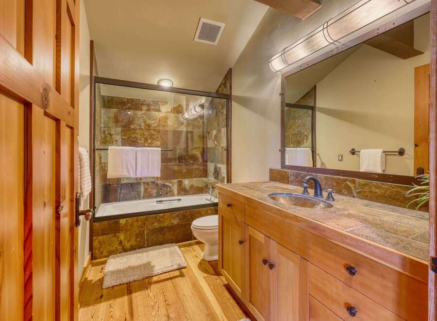 This rustic bathroom features a wooden vanity with a stone countertop and a stone-tiled shower/tub surround.