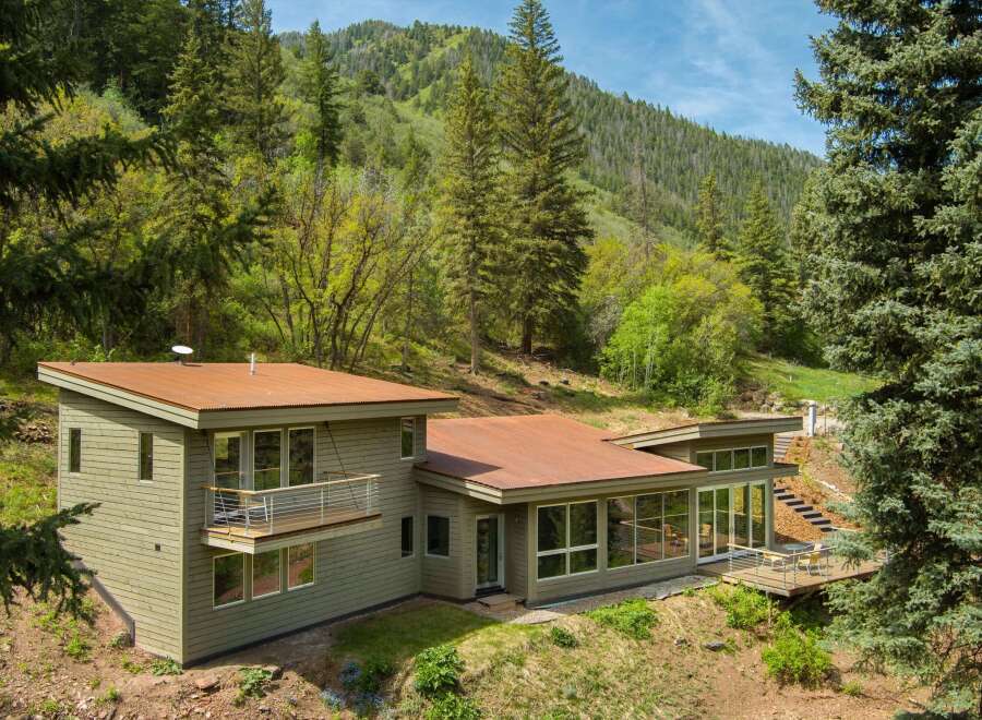 Nestled amidst a lush forest, this modern home boasts a striking design with a rusted metal roof and expansive windows that invite the natural surroundings indoors.