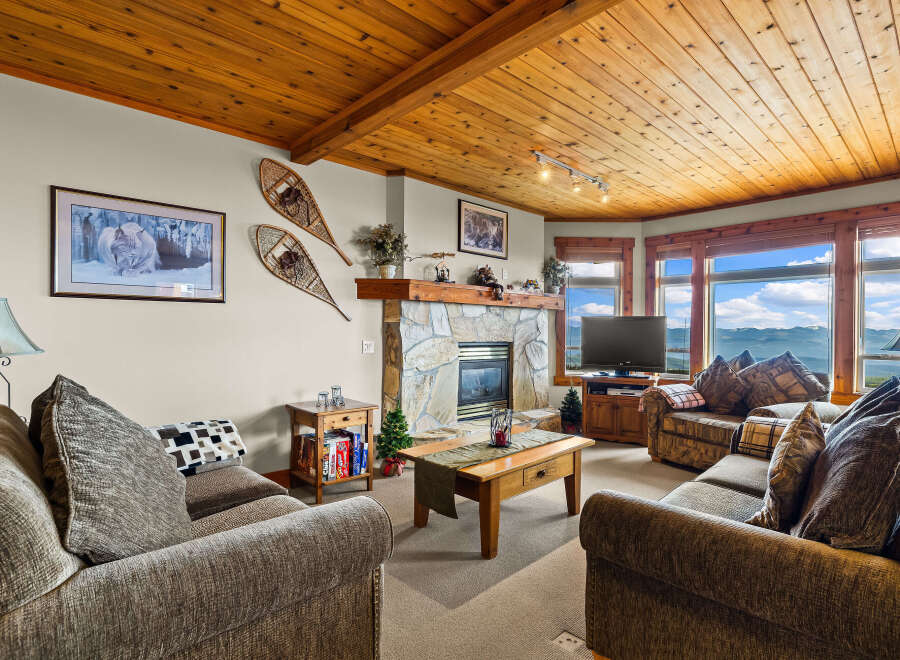 Experience breathtaking mountain views from this cozy living room, featuring a stone fireplace and a warm, wood-paneled ceiling.
