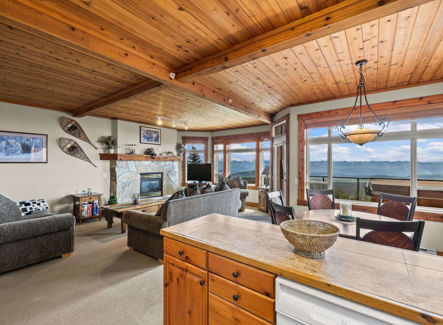 Experience breathtaking mountain views from this stunning living and dining area, featuring a cozy stone fireplace and a warm, wood-paneled ceiling.