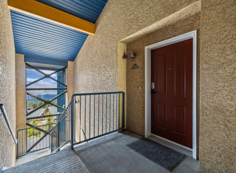 Experience mountain living with this charming condo's exterior entrance, featuring a striking red door and a scenic balcony overlooking a wooded landscape.