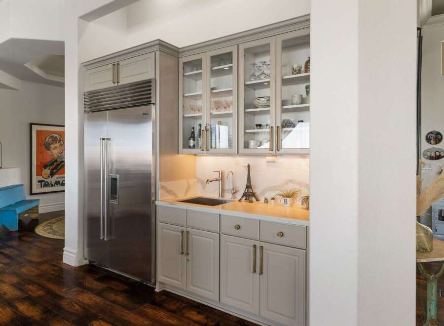 This wet bar area features a sleek stainless steel refrigerator and ample storage with grey cabinetry and glass-fronted upper cabinets displaying glassware and decor.