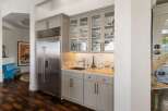 This wet bar area features a sleek stainless steel refrigerator and ample storage with grey cabinetry and glass-fronted upper cabinets displaying glassware and decor.