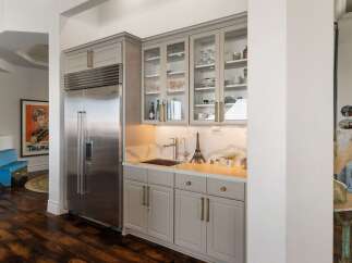 This wet bar area features a sleek stainless steel refrigerator and ample storage with grey cabinetry and glass-fronted upper cabinets displaying glassware and decor.