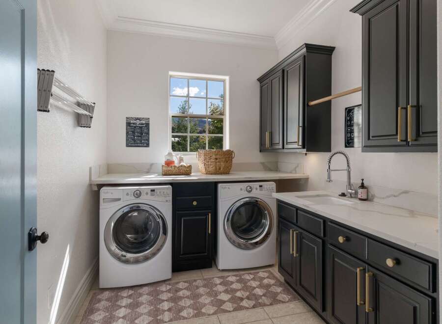 This spacious laundry room features modern dark cabinetry with elegant gold hardware, complemented by a sleek white countertop and a deep sink.