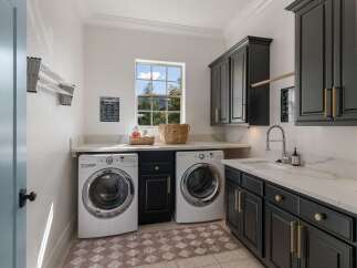 This spacious laundry room features modern dark cabinetry with elegant gold hardware, complemented by a sleek white countertop and a deep sink.