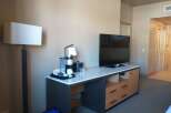 This hotel room features a modern desk area with a coffee maker, mini-fridge, and a flat-screen TV.