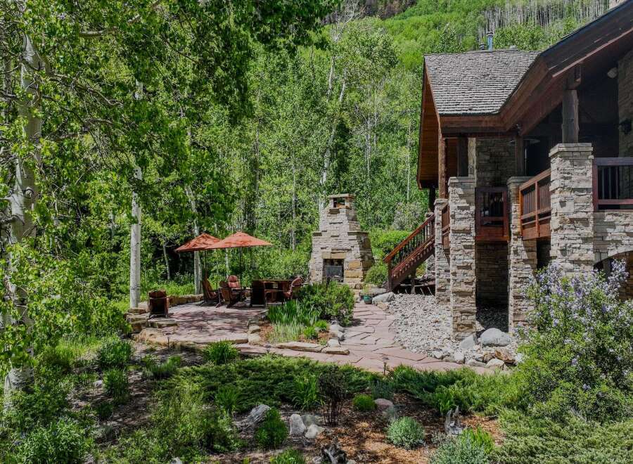 Enjoy the serene mountain lifestyle with this stunning outdoor living space, featuring a charming stone fireplace and comfortable seating under two large umbrellas.