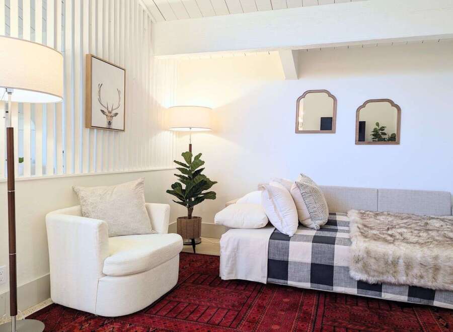This cozy living space features a comfortable white armchair and a stylish daybed adorned with a buffalo check blanket and a plush faux fur throw.